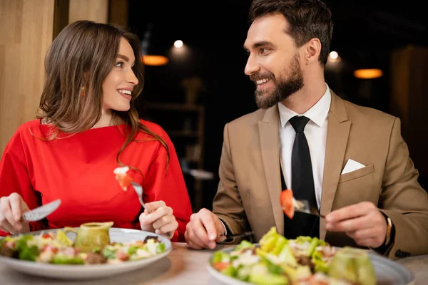 Feliz joven pareja en traje festivo mirándose en el restaurante durante la celebración en el día de San Valentín - foto de stock