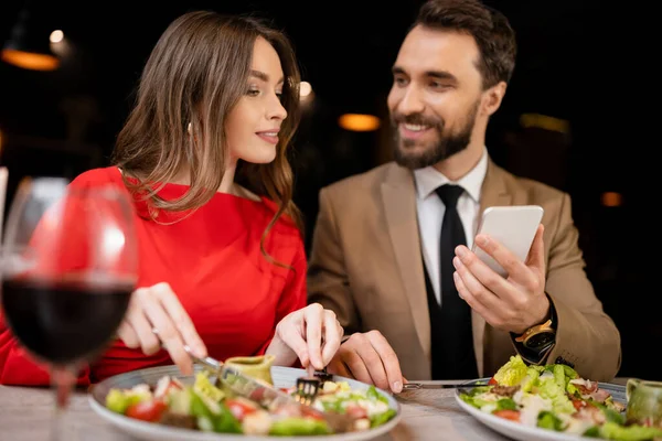 Hombre barbudo usando teléfono inteligente cerca de novia feliz en el restaurante durante la celebración en el día de San Valentín - foto de stock