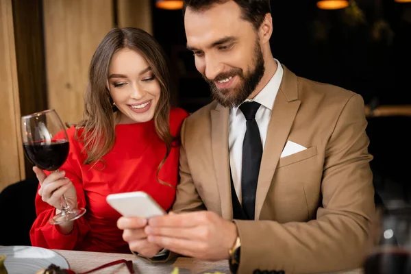 Бородатий чоловік використовує смартфон поруч із щасливою дівчиною з келихом вина в ресторані — стокове фото
