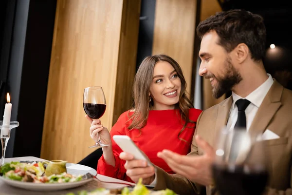 Bearded man holding smartphone near happy girlfriend with glass of wine during celebration on valentines day — Stock Photo