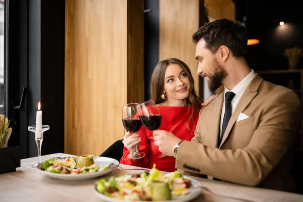 Cheerful young woman and bearded man clinking glasses with red wine during celebration on valentines day — Stock Photo
