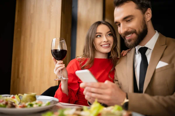 Hombre alegre sosteniendo teléfono inteligente cerca de novia feliz con copa de vino durante la celebración en el día de San Valentín - foto de stock