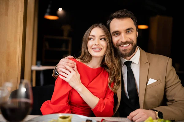 Bearded man hugging cheerful young woman in red dress and looking at camera on valentines day — Stock Photo