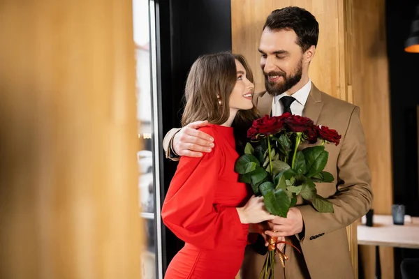 Feliz hombre sosteniendo rosas rojas mientras abraza alegre novia en vestido en el día de San Valentín - foto de stock