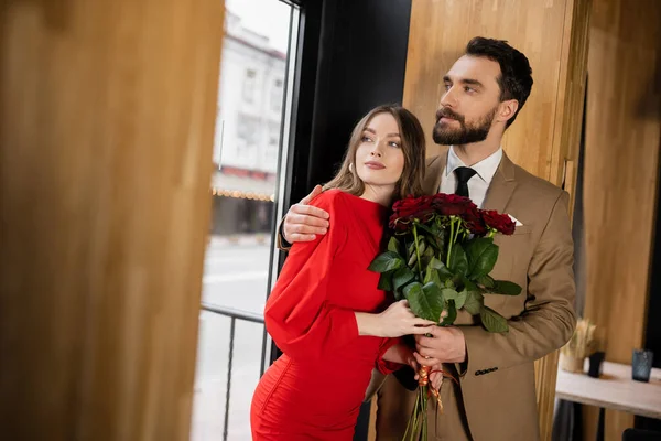 Barbudo homem segurando rosas vermelhas e abraçando jovem mulher no vestido no dia dos namorados — Fotografia de Stock