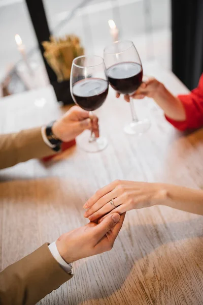 Vista recortada de la mujer con anillo de compromiso en el dedo tintineo vasos de vino con novio en el día de San Valentín - foto de stock