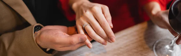 Vista recortada del hombre sosteniendo la mano de la mujer con anillo de compromiso en el dedo en el día de San Valentín, bandera - foto de stock