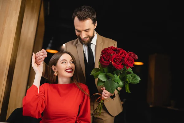 Hombre barbudo en ropa formal celebración ramo de rosas rojas cerca de mujer alegre en el día de San Valentín - foto de stock