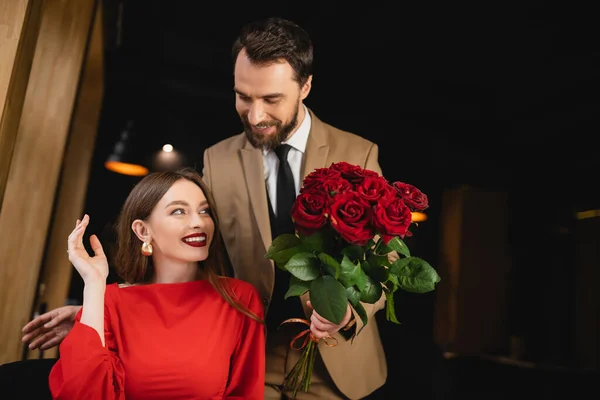 Bearded man in formal wear holding bouquet of red roses near happy woman on valentines day — Stock Photo