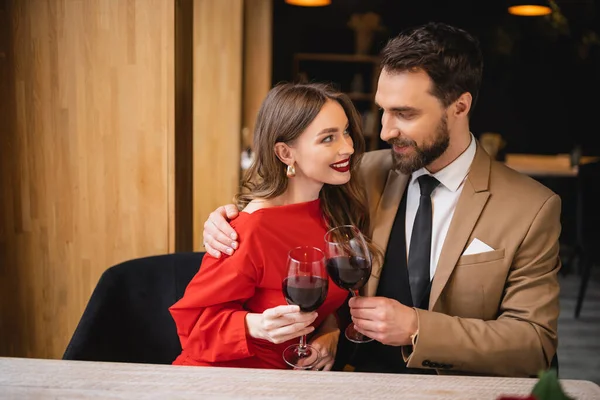 Mujer positiva tintineo vasos de vino con barbudo hombre en el día de San Valentín - foto de stock