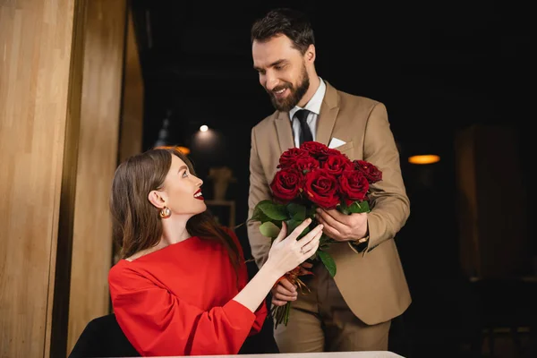 Hombre barbudo en ropa formal que sostiene ramo de rosas rojas cerca de la mujer complacida en el día de San Valentín - foto de stock