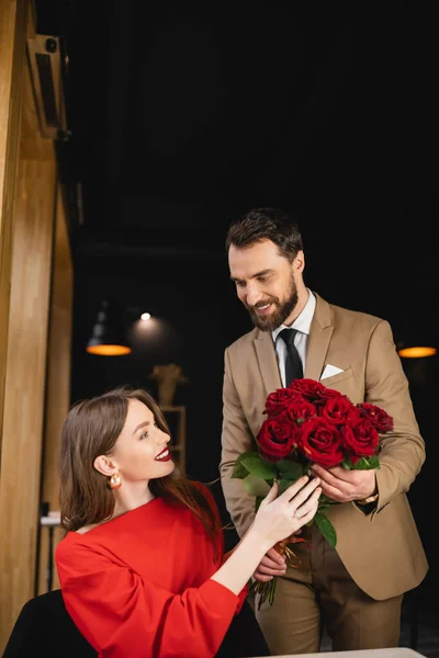 Bearded man in formal wear holding bouquet of red roses near smiling woman on valentines day — Stock Photo