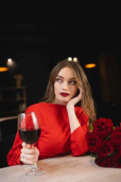 Joven soñadora sosteniendo copa de vino tinto cerca de rosas en el día de San Valentín - foto de stock
