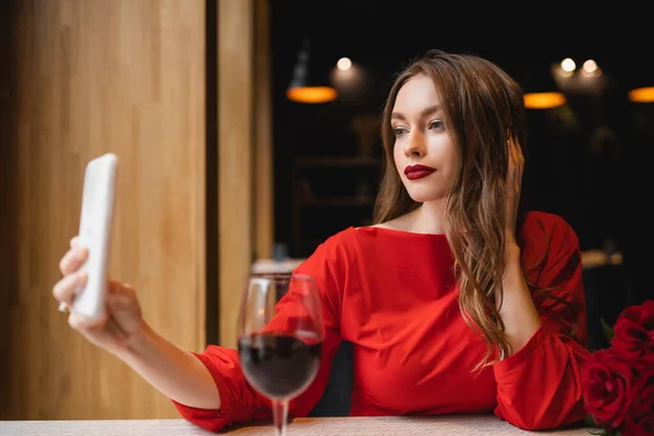 Pretty young woman with red lips taking selfie near glass of wine and roses on valentines day — Stock Photo