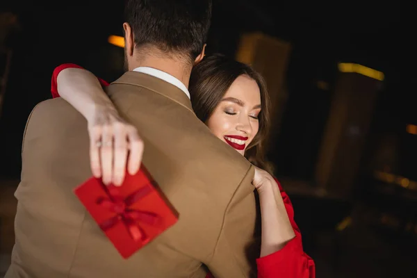 Весела жінка з закритими очима тримає подарунок і обіймає хлопця в день валентинки — стокове фото