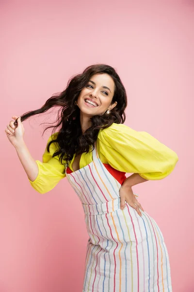 Coquettish housewife in bright yellow blouse and apron holding wavy hair and posing with hand on hip isolated on pink — Stock Photo