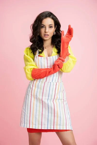 Morena ama de casa en blusa amarilla y delantal rayado con guante de goma roja y mirando a la cámara aislada en rosa - foto de stock