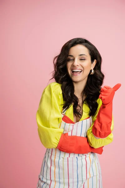 Excited brunette housewife in red rubber gloves laughing and pointing with thumb isolated on pink — Stock Photo
