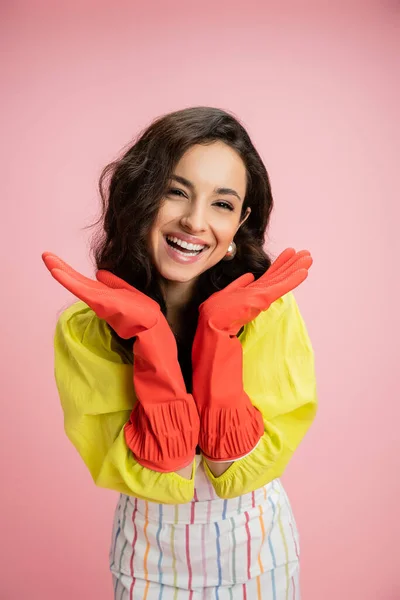 Morena complacida ama de casa en blusa amarilla y guantes de goma rojos tomados de la mano cerca de la cara aislada en rosa - foto de stock