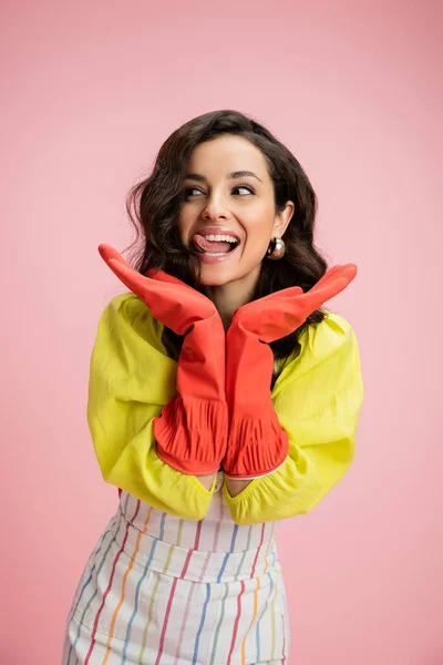Excited housewife in yellow blouse and red rubber gloves holding hands near face and sticking out tongue isolated on pink — Stock Photo