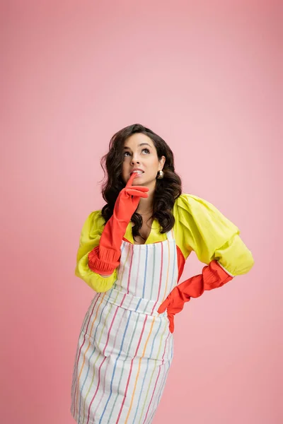 Thoughtful housewife in striped apron and red rubber gloves touching lips and looking up isolated on pink — Stock Photo