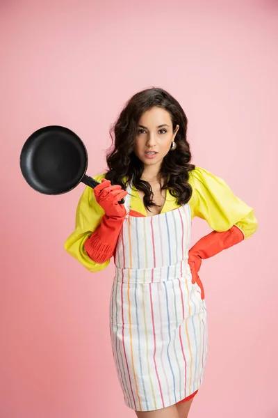 Brunette housewife in red rubber gloves and striped apron standing with frying pan and hand on hip isolated on pink — Stock Photo