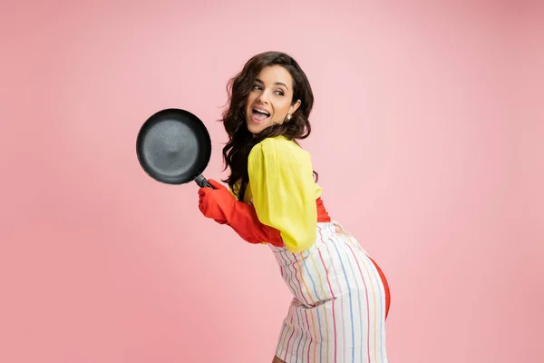 Flirty and astonished housewife in yellow blouse holding frying pan and looking away isolated on pink — Stock Photo