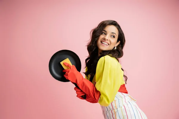 Cheerful and stylish housewife with frying pan and sponge looking away isolated on pink — Stock Photo
