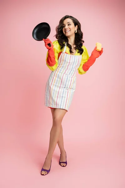 Full length of cheerful brunette housewife in striped apron posing with sponge and frying pan on pink — Stock Photo