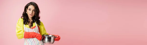 Jovem e bonita dona de casa segurando panela e olhando para a câmera isolada em rosa, banner — Fotografia de Stock