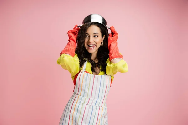 Flirty housewife in red rubber gloves holding saucepan above head and sticking out tongue isolated on pink — Stock Photo