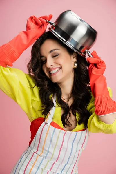 Cheerful woman in red rubber gloves having fun while holding saucepan above head isolated on pink — Stock Photo