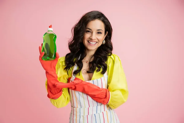 Brunette housewife in red rubber gloves showing dishwashing liquid and smiling at camera isolated on pink — Stock Photo