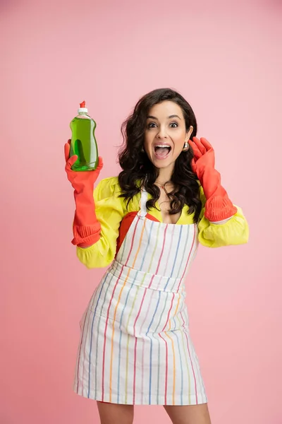 Sorprendido ama de casa en guantes de goma roja y delantal rayado que sostiene el líquido de lavado de platos aislado en rosa - foto de stock