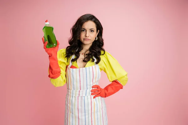 Displeased and frowning housewife in red rubber gloves holding dishwashing liquid while standing with hand on hip isolated on pink — Stock Photo