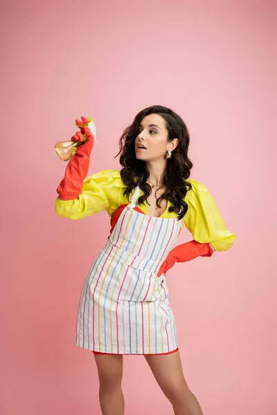 Bruna casalinga in camicetta gialla e grembiule a righe con bottiglia spray e guardando lontano isolato sul rosa — Foto stock