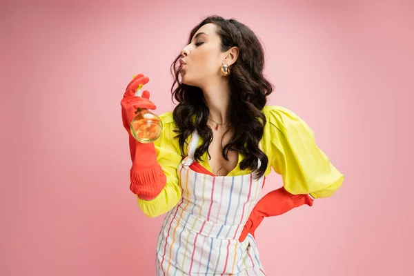 Woman in striped apron and red rubber gloves blowing at spray bottle isolated on pink — Stock Photo