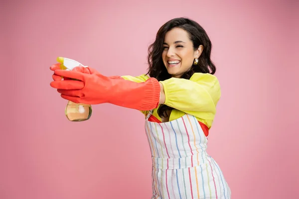 Cheerful housewife in striped apron and red rubber gloves having fun and aiming with spray bottle isolated on pink — Stock Photo