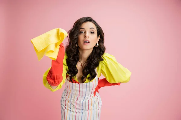 Young housewife in striped apron and red rubber gloves holding yellow rag and looking at camera isolated on pink — Stock Photo