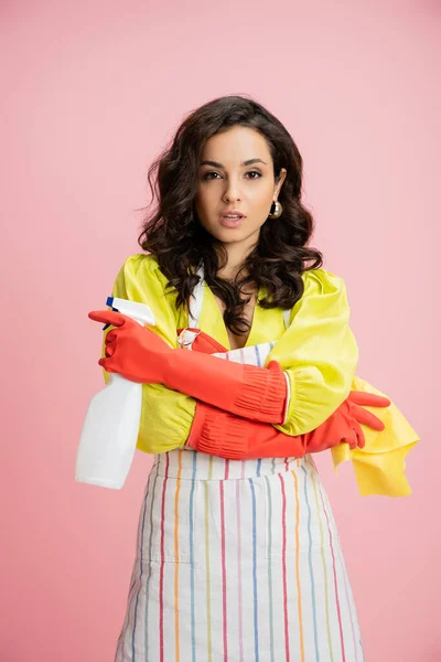 Stylish brunette housewife in red rubber gloves standing with rag and spray bottle in crossed arms isolated on pink — Stock Photo