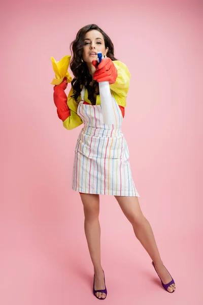 Full length of woman in striped apron and rubber gloves holding spray bottle and rag while looking at camera on pink — Stock Photo