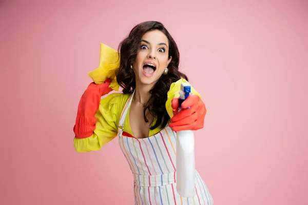 Excited housewife having fun while holding rag and spraying from bottle at camera isolated on pink — Stock Photo