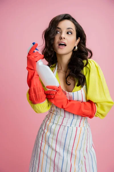 Curiosa ama de casa en guantes de goma roja y blusa amarilla sosteniendo botella de spray y mirando hacia otro lado aislado en rosa - foto de stock