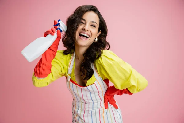 Lachende Hausfrau in gelber Bluse und roten Gummihandschuhen mit Sprühflasche und posiert mit der Hand an der Hüfte isoliert auf rosa — Stockfoto