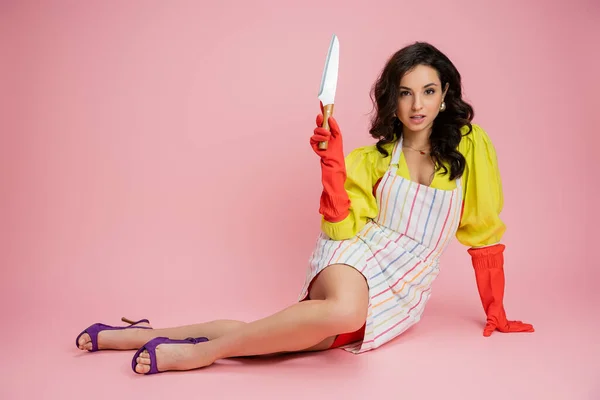 Longitud completa de la elegante ama de casa en blusa amarilla y delantal a rayas sentado con cuchillo de cocina y mirando a la cámara en el fondo rosa - foto de stock