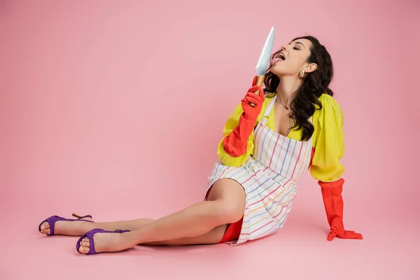 Longitud completa de ama de casa sensual en guantes de goma roja lamiendo cuchillo de cocina mientras está sentado sobre fondo rosa - foto de stock