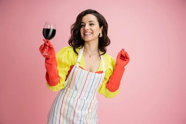 Coquete dona de casa em luvas de borracha vermelha e avental sorrindo e segurando copo de vinho isolado em rosa — Fotografia de Stock