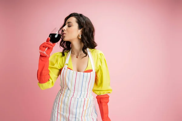 Dona de casa morena em blusa amarela e avental listrado com luvas de borracha vermelha bebendo vinho isolado em rosa — Fotografia de Stock