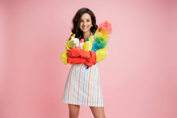 Joyeuse jeune femme au foyer avec brosse à poussière colorée et des fournitures de nettoyage en regardant la caméra isolée sur rose — Photo de stock