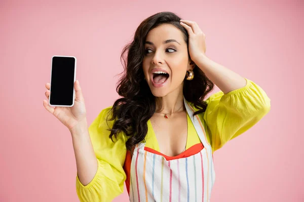 Astonished woman in yellow blouse and striped apron touching head while holding cellphone with blank screen isolated on pink — Stock Photo